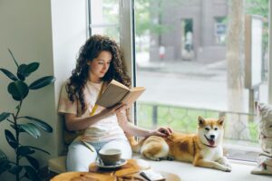 woman reading a book near a windowsill with her dog